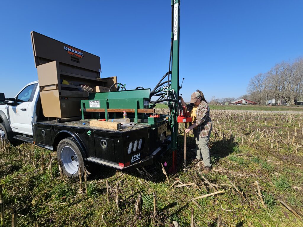The picture features Amber collecting deep soil cores using a hydraulic probe attached to the back of a truck.