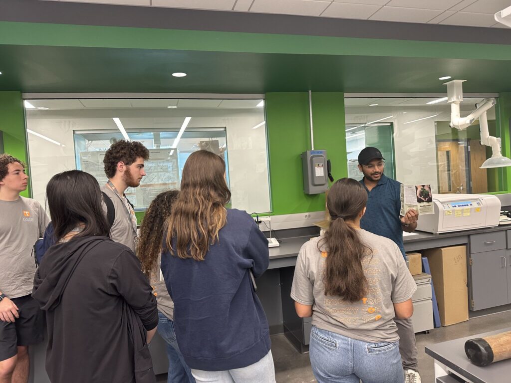 The photo features Dr. Neelipally explaining the concepts of soil health to high school students with handouts.