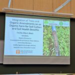 The photo presents the title of Cecilia's defense presentation on the projector as "Integration of trees and organic amendments on an organic farm for soil carbon and soil health benefits."